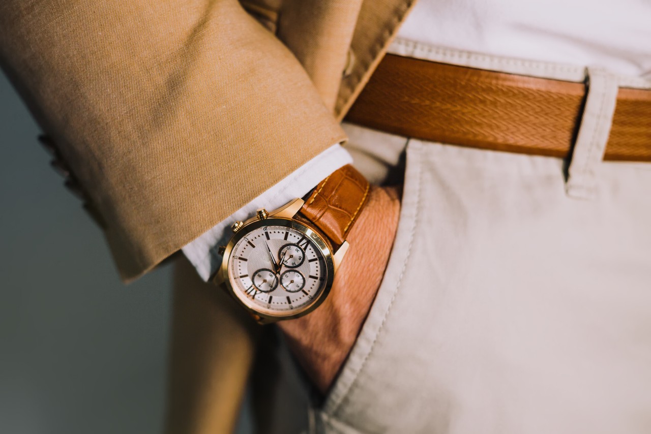 A close up of man’s hand in the pocket of his white trousers, showcasing a brown leather watch that matches his belt.