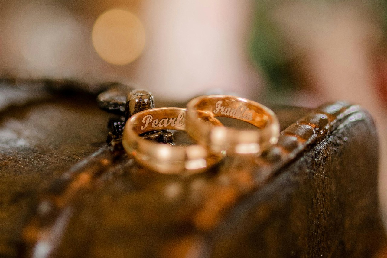 A pair of yellow gold wedding bands atop a leather chest, with the names “Pearl” and “Franklin” engraved inside.