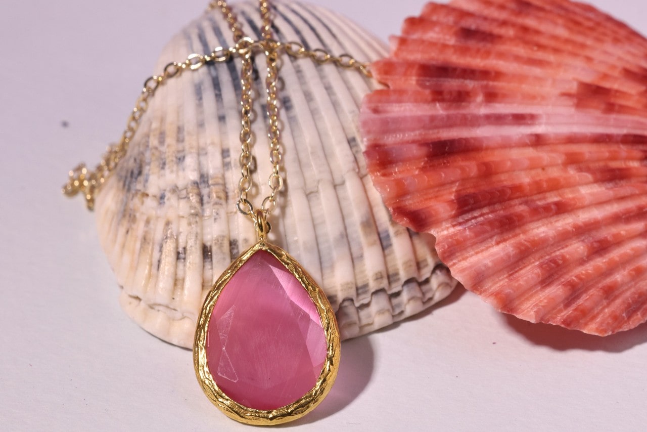 a yellow gold pendant necklace featuring a pink gemstone lying over two shells a yellow gold pendant necklace featuring a pink gemstone lying over two shells