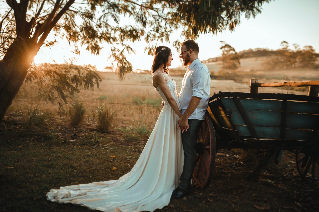 A newlywed couple stands under a tree during sunset, holding hands and gazing into each other's eyes. The bride wears a flowing white gown, while the groom wears a white shirt and dark trousers. A newlywed couple stands under a tree during sunset, holding hands and gazing into each other's eyes. The bride wears a flowing white gown, while the groom wears a white shirt and dark trousers.