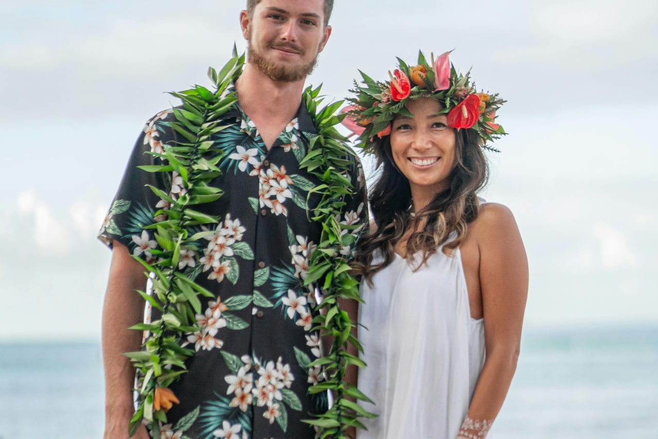A smiling couple dressed in Hawaiian attire stands by the ocean, with the man wearing a leafy lei over a floral shirt and the woman adorned with a vibrant flower crown and a flowing white dress. A smiling couple dressed in Hawaiian attire stands by the ocean, with the man wearing a leafy lei over a floral shirt and the woman adorned with a vibrant flower crown and a flowing white dress.
