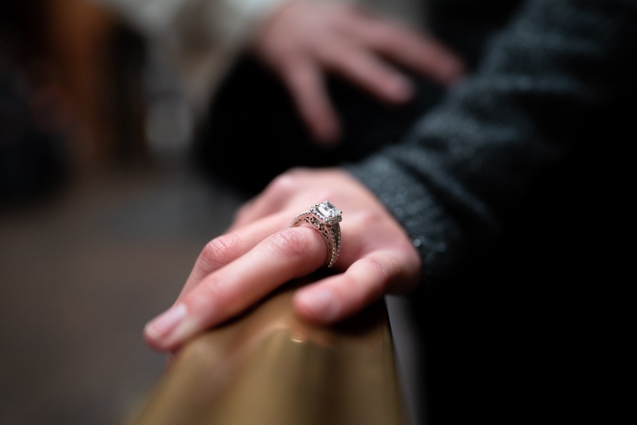 a closeup of a woman’s hand wearing an engagement ring resting on the back of a pew.