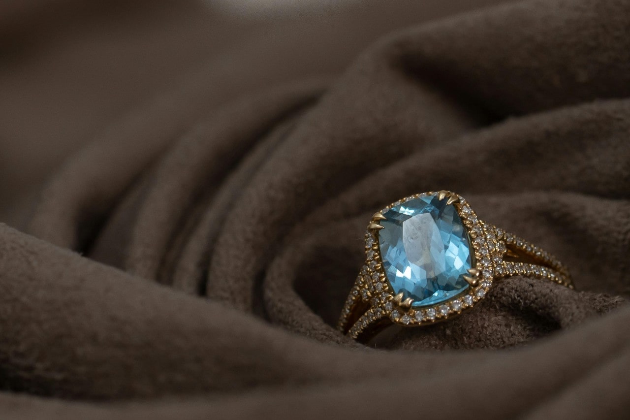 A luxurious gold ring featuring a large, cushion-cut blue gemstone, surrounded by a halo of sparkling diamonds, elegantly placed on soft brown fabric.